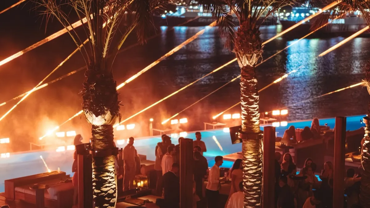 Luxury Mykonos nightlife scene at a waterfront beach club with illuminated palm trees, guests, poolside seating and dramatic evening lighting.