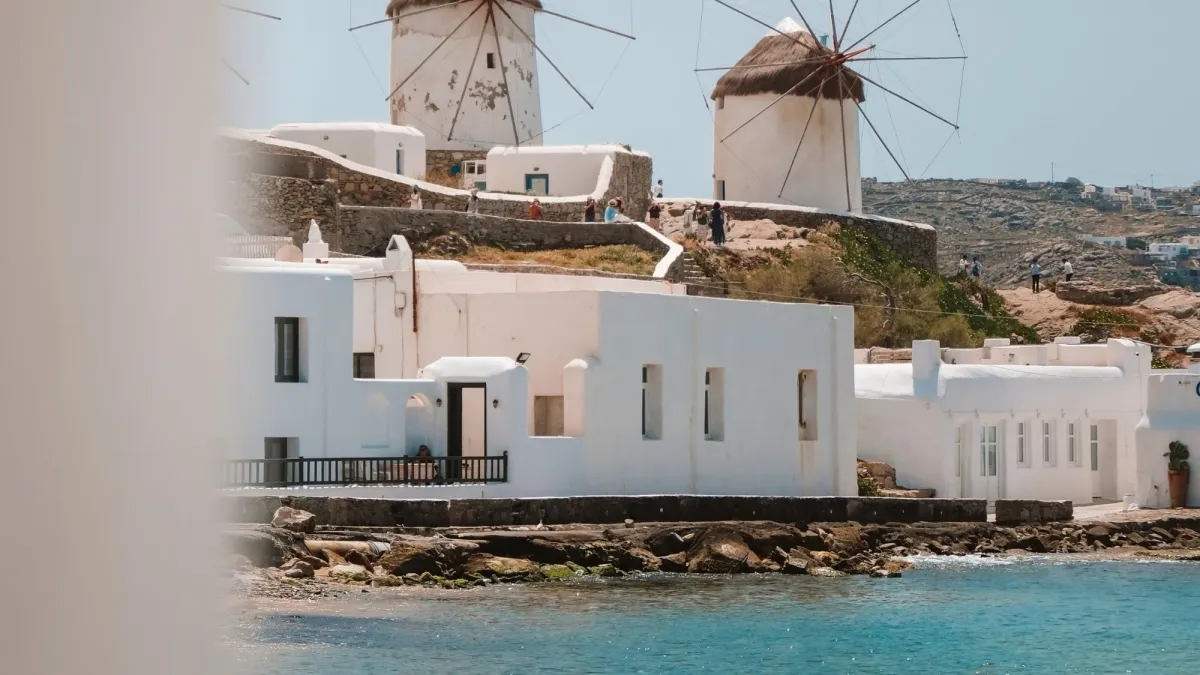 Traditional Mykonos windmills rising above whitewashed Cycladic houses beside the blue Aegean Sea, capturing the iconic landscape of Mykonos Town, Greece.