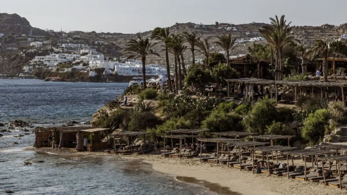 Luxury beachfront setting in Mykonos with sunbeds, palm trees and traditional white Cycladic buildings overlooking the Aegean Sea.