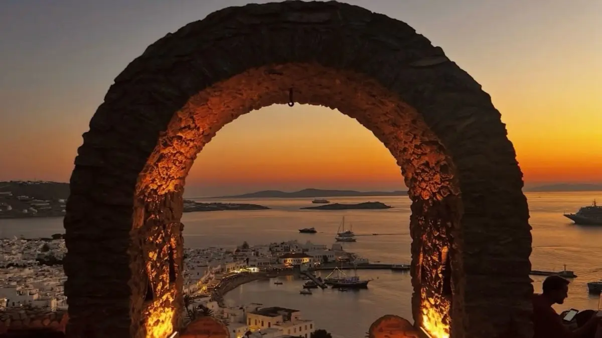 Sunset view overlooking Mykonos Town and the Aegean Sea through a stone archway with warm evening lighting