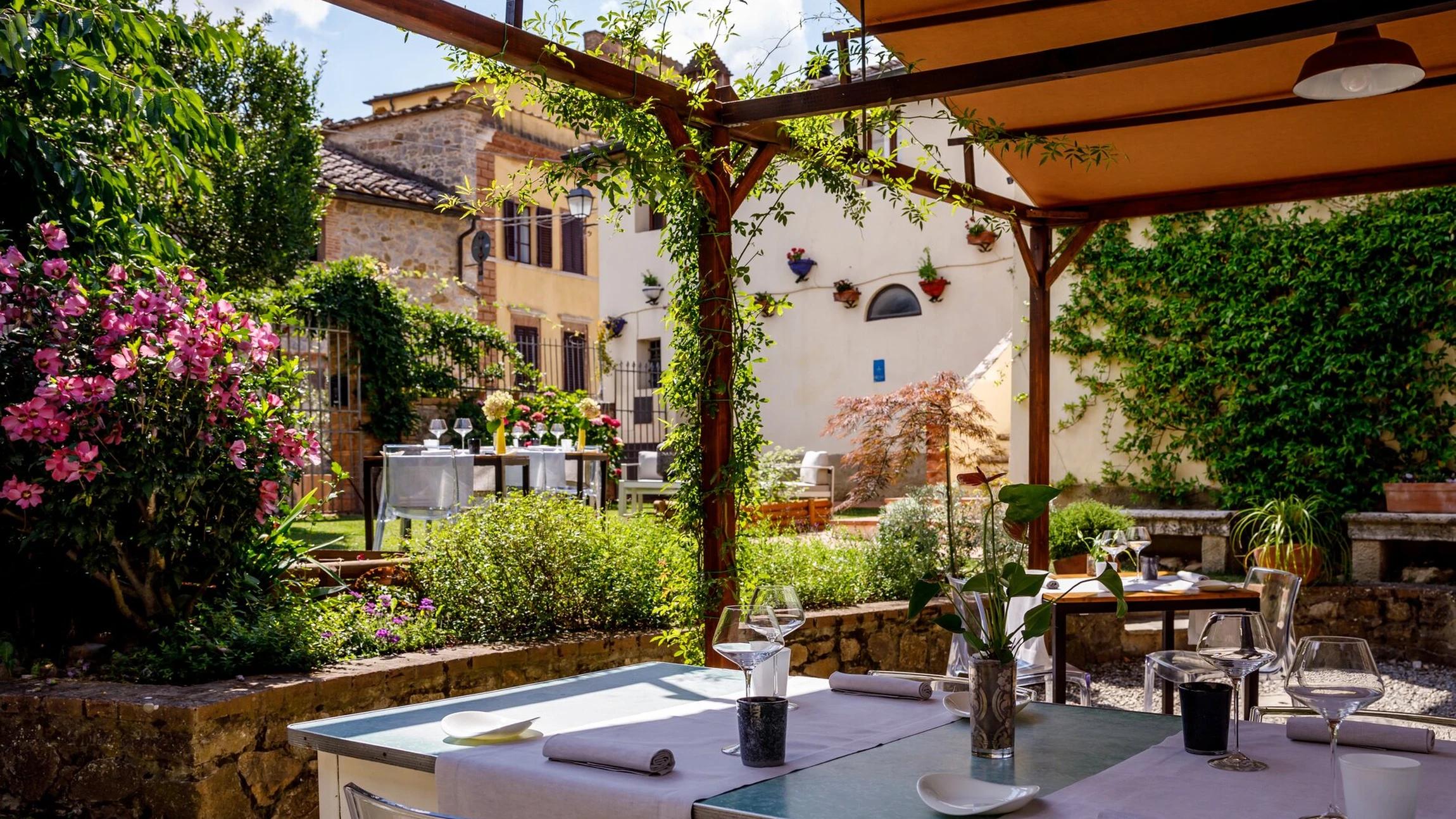 The garden at L’Asinello in Villa a Sesta: tables laid under a vine-covered pergola, with flowering hibiscus and the village houses behind