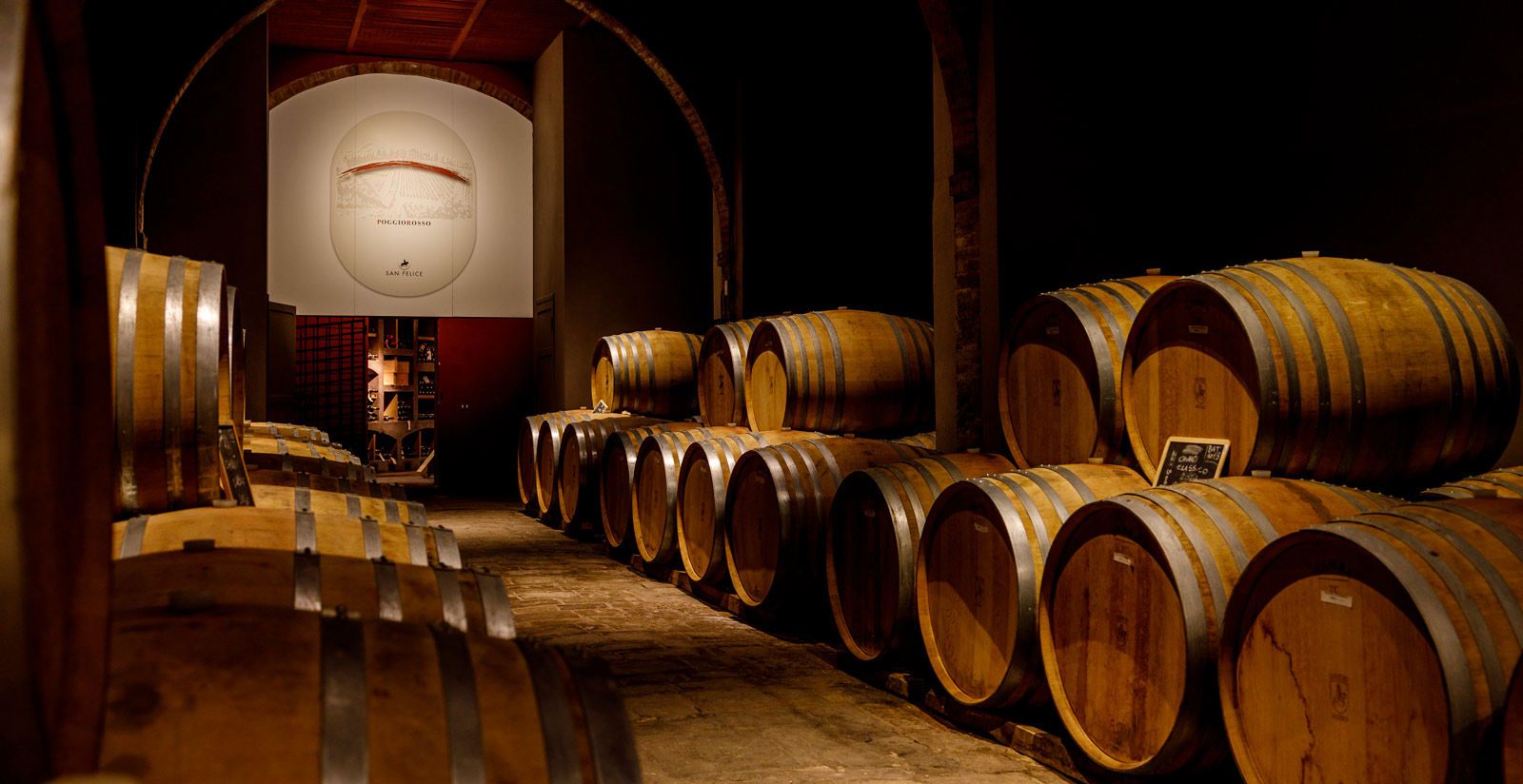The barrel cellar at San Felice: rows of oak barriques beneath brick arches, with the Poggio Rosso emblem on the far wall