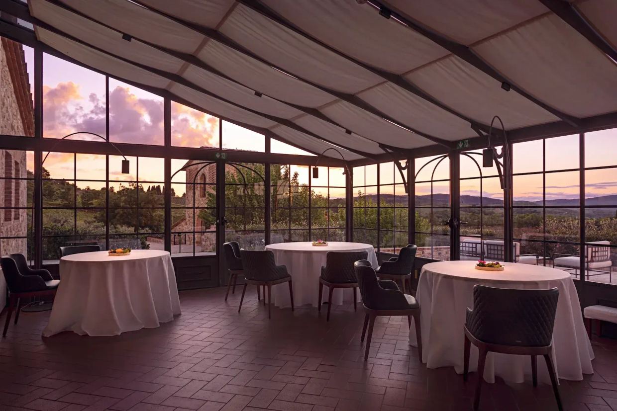 The dining room at Il Visibìlio at sunset: round tables laid in white inside a glass-and-iron conservatory, with the hills beyond