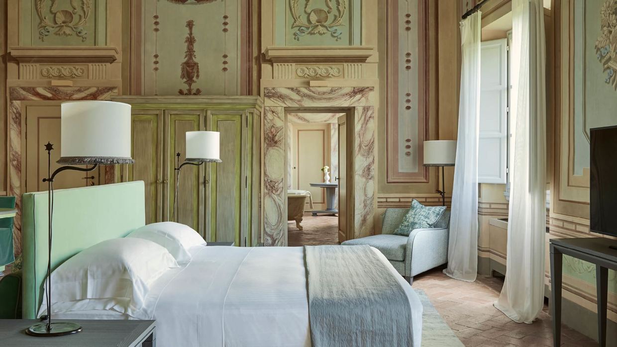 A suite at COMO Castello Del Nero: frescoed walls and painted-marble door frames around a white bed, with a freestanding bath glimpsed in the next room