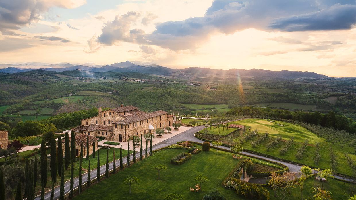 Aerial view of The Club House at Pievasciata at sunset: a long stone building with a cypress-lined drive, lawns and a golf green, hills fading into the distance