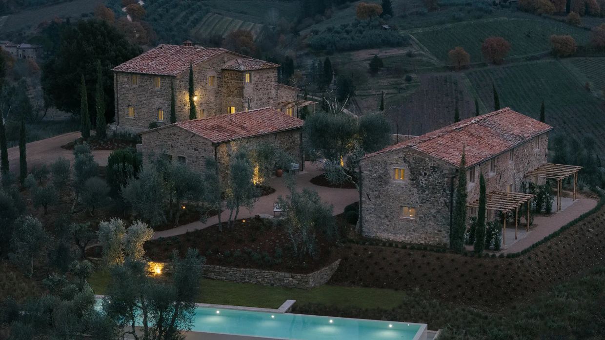 La Darbia in Chianti at dusk: three stone farmhouses with lit windows among olive trees and cypresses, the pool in the foreground and vineyards on the hillside behind
