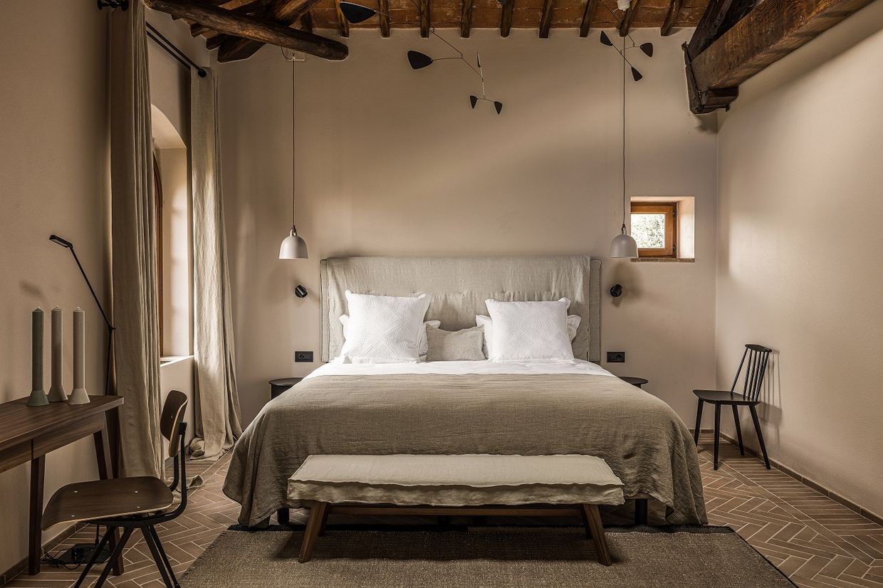 A bedroom at Pieve Aldina: linen bed and bench under old wooden beams, with herringbone terracotta floors and soft natural tones