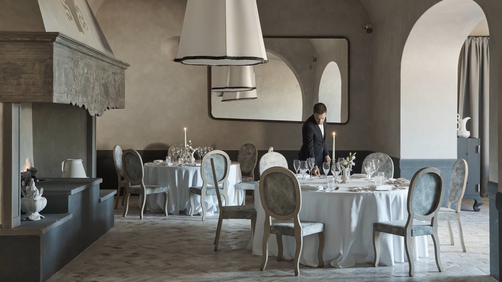 The dining room at La Torre, COMO Castello Del Nero: candlelit round tables beneath vaulted arches, with a carved stone fireplace and a waiter setting a table