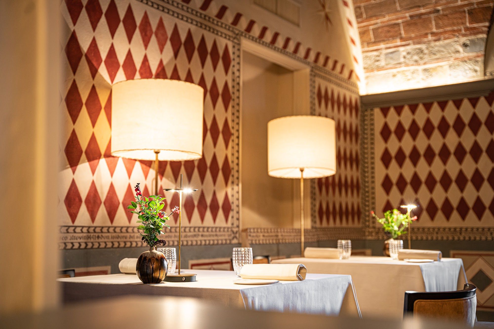The dining room at Osteria di Passignano: tables lit by tall lamps beneath a brick vault, with walls painted in red diamonds and a frieze of names and dates