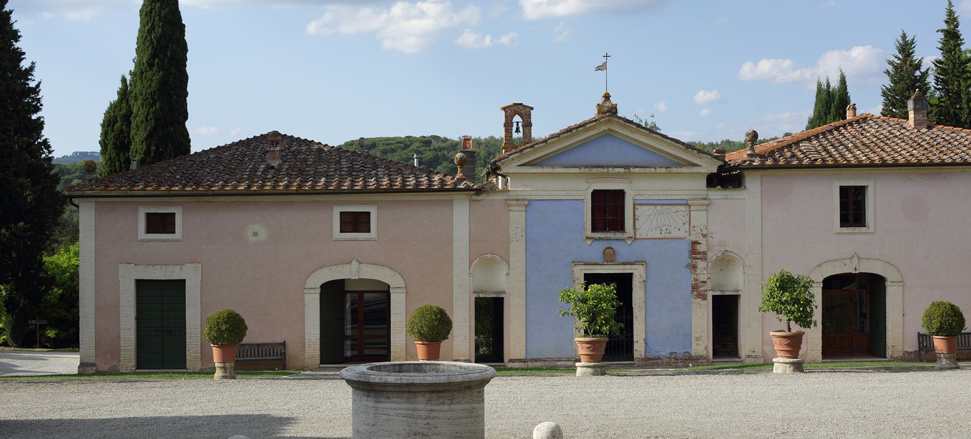 The courtyard at Fèlsina: pink and blue façades around a small chapel with a bell gable and sundial, with potted lemon trees and an old stone well in front