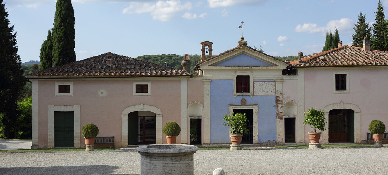 The courtyard at Fèlsina: pink and blue façades around a small chapel with a bell gable and sundial, with potted lemon trees and an old stone well in front