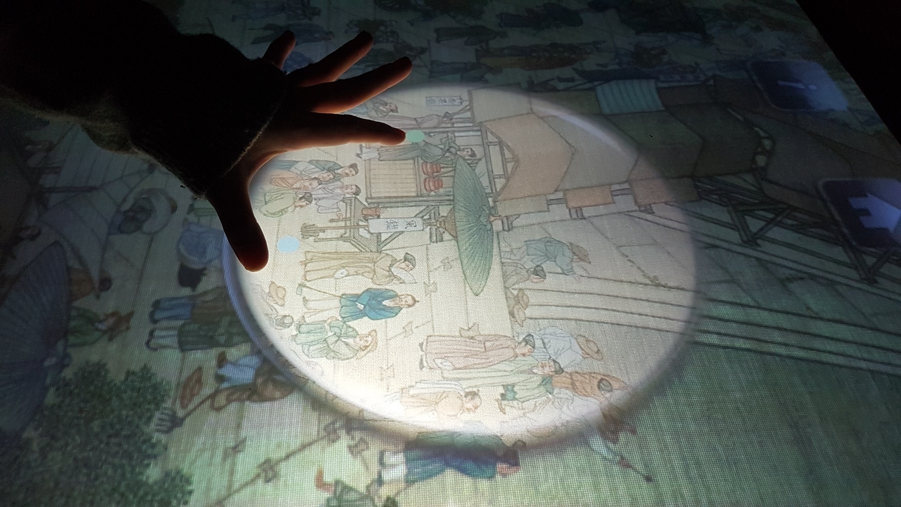 手放在互動桌面上，光圈放大畫中細節。/ A hand on an interactive table, a circle of light magnifying details of the painting.
