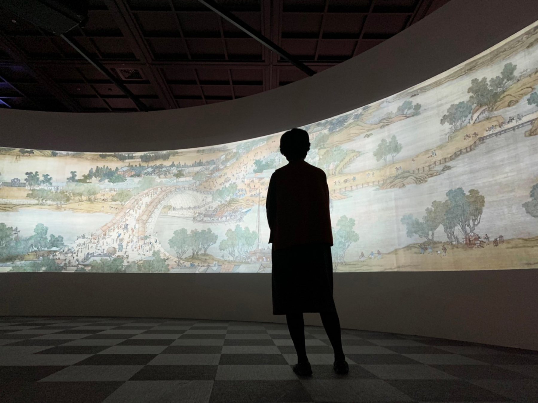 觀眾剪影站在清明上河圖弧形投影牆前。/ A visitor in silhouette before a curved projection of the scroll.