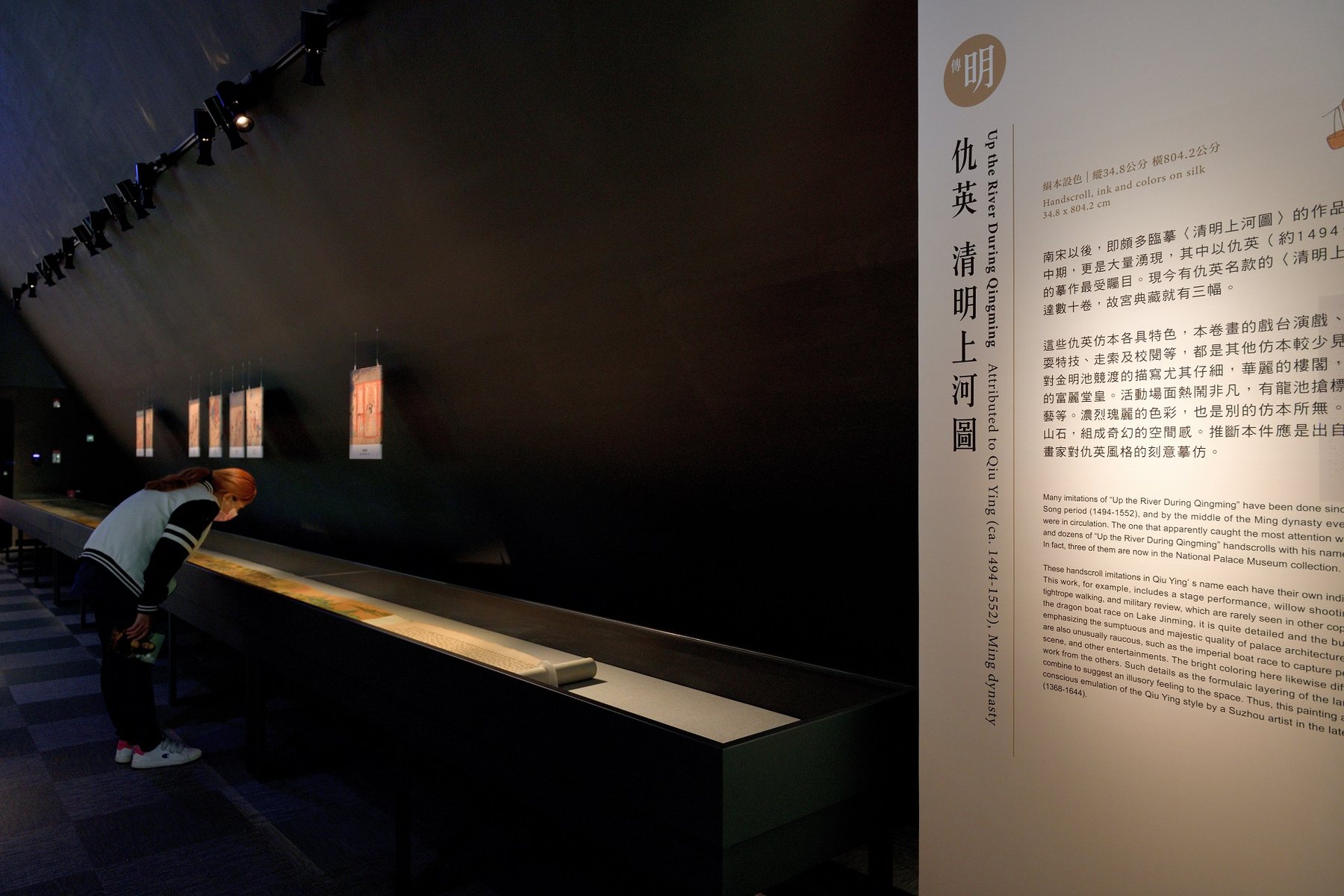 觀眾俯看長卷展櫃，旁為仇英《清明上河圖》說明牌。/ A visitor leaning over the scroll showcase beside the label for Qiu Ying’s version.