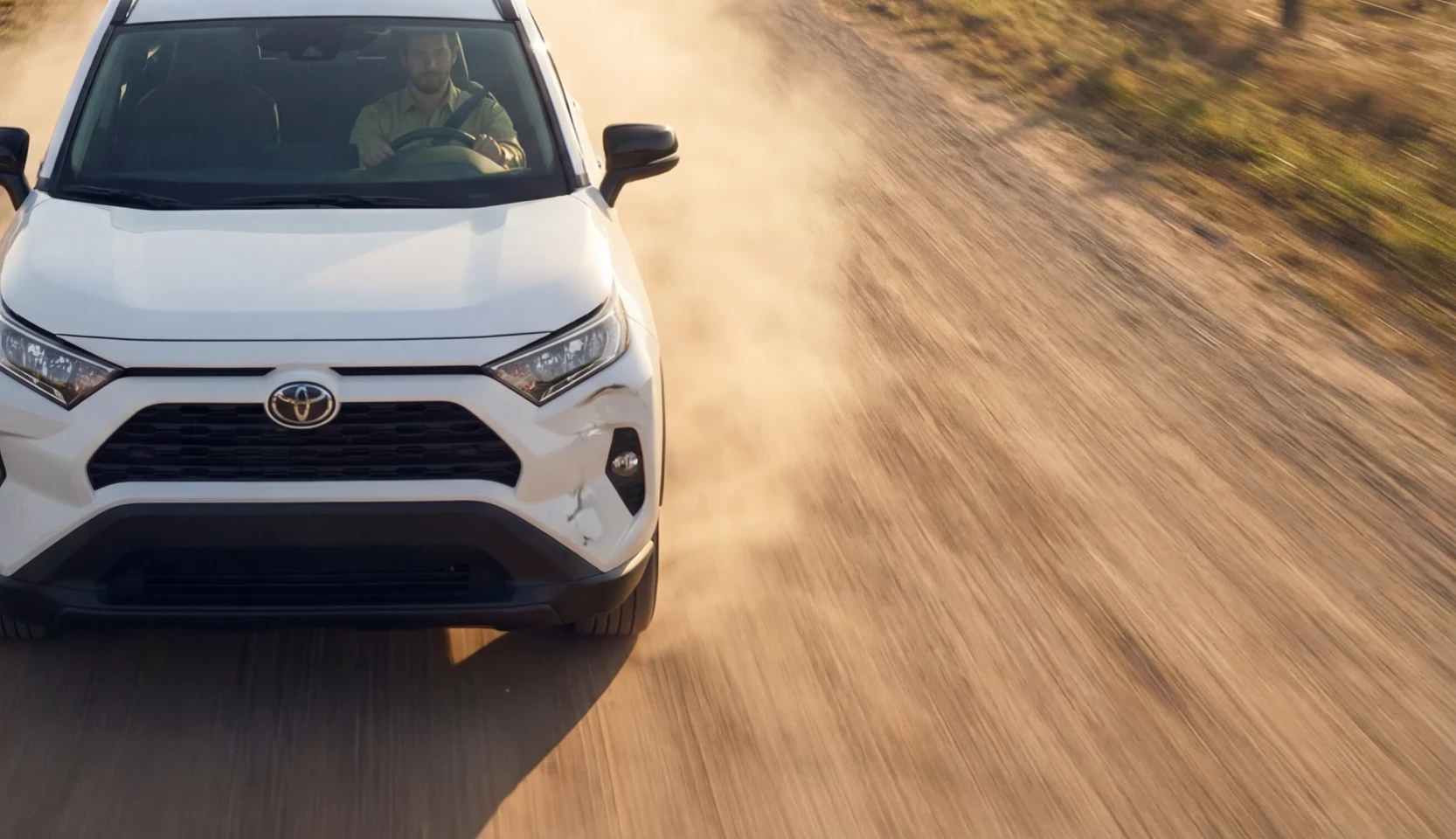 White Toyota RAV4 driving on a dusty road, with a dent on the front bumper below the right headlight