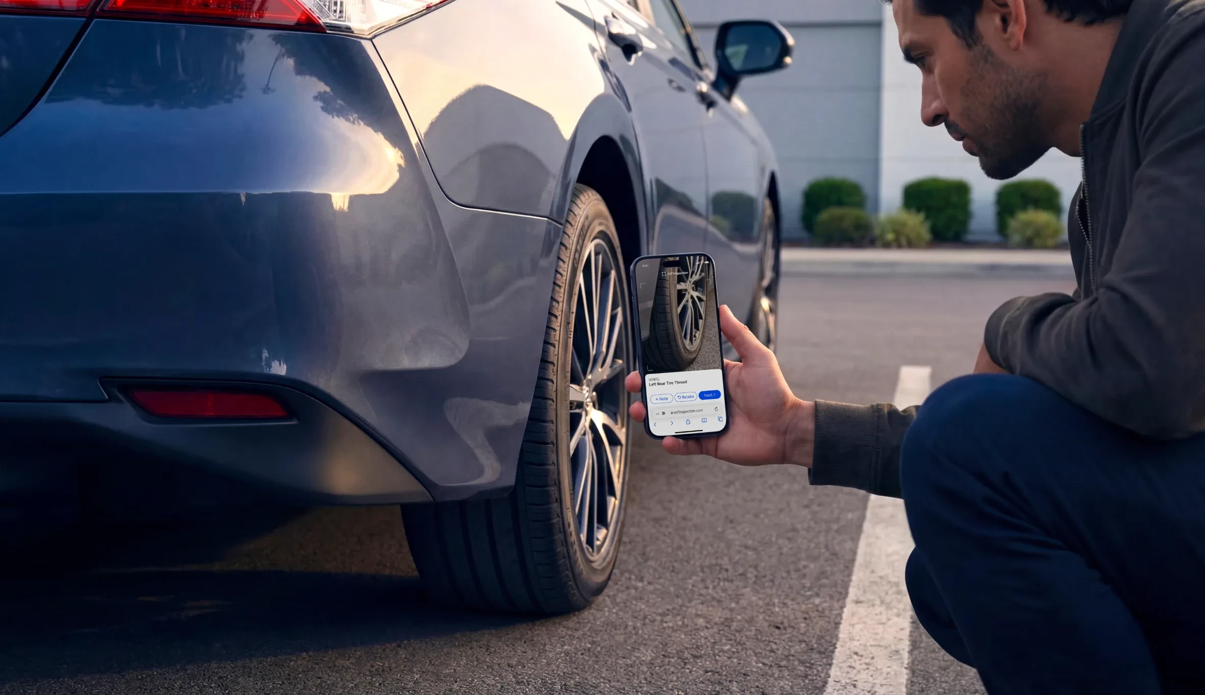 Man crouched beside a sedan capturing the left rear tire tread with a phone