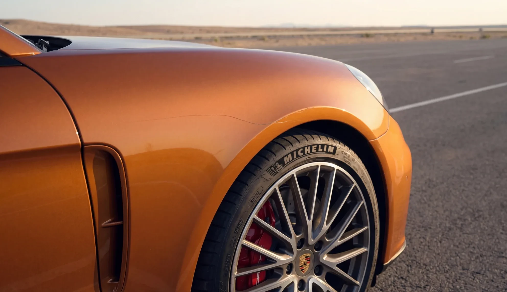 Front fender and wheel of an orange Porsche in a desert parking lot, with a scratch along the wheel arch