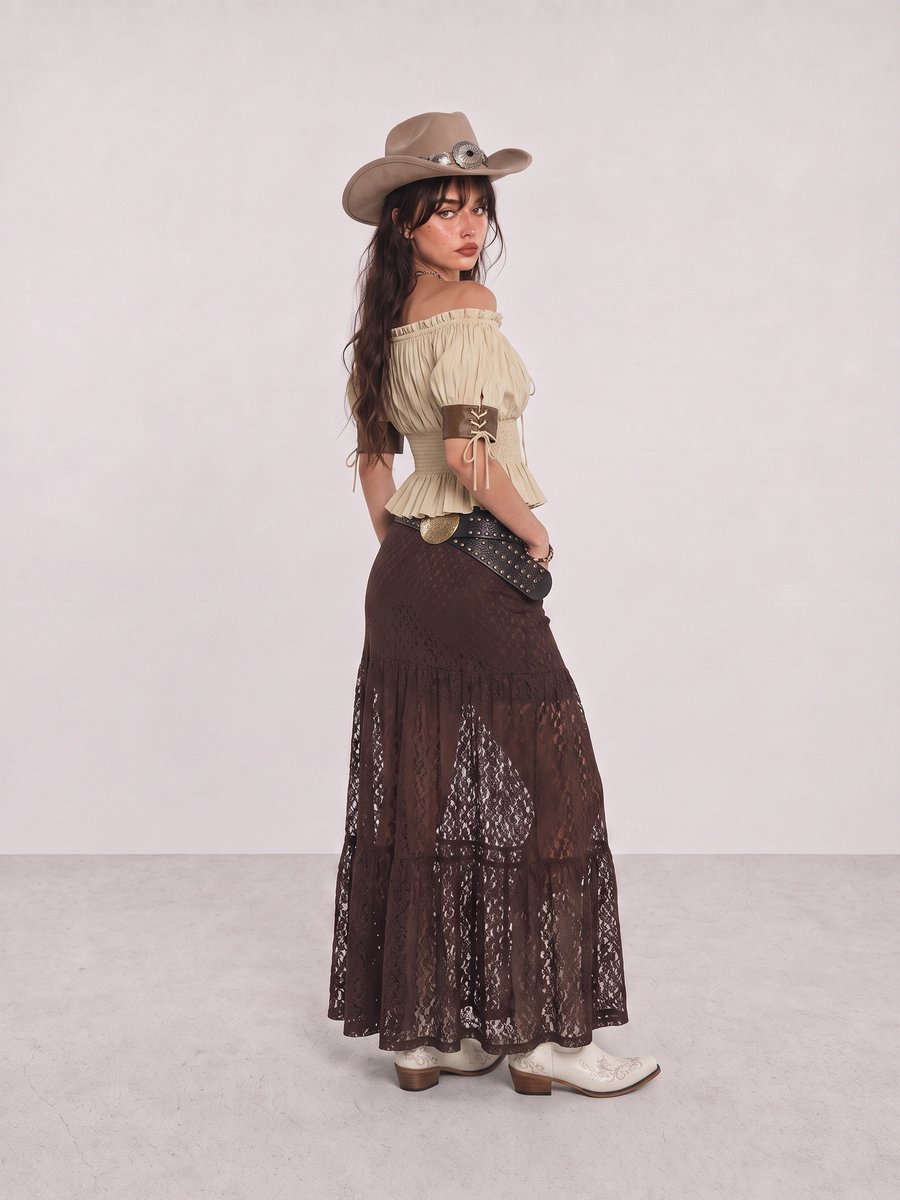 Woman wearing a beige fringe top, brown lace maxi skirt, white cowboy boots, and a taupe hat
