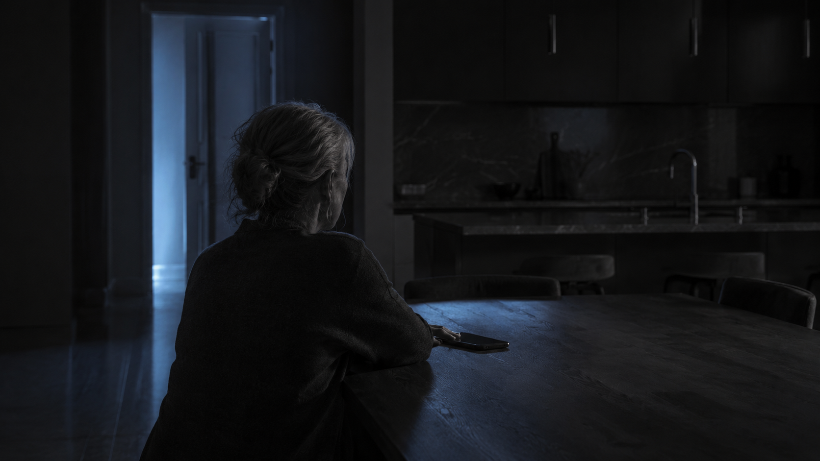 Person alone at a kitchen table at night after noticing signs of an emotional affair, phone face down beside them.