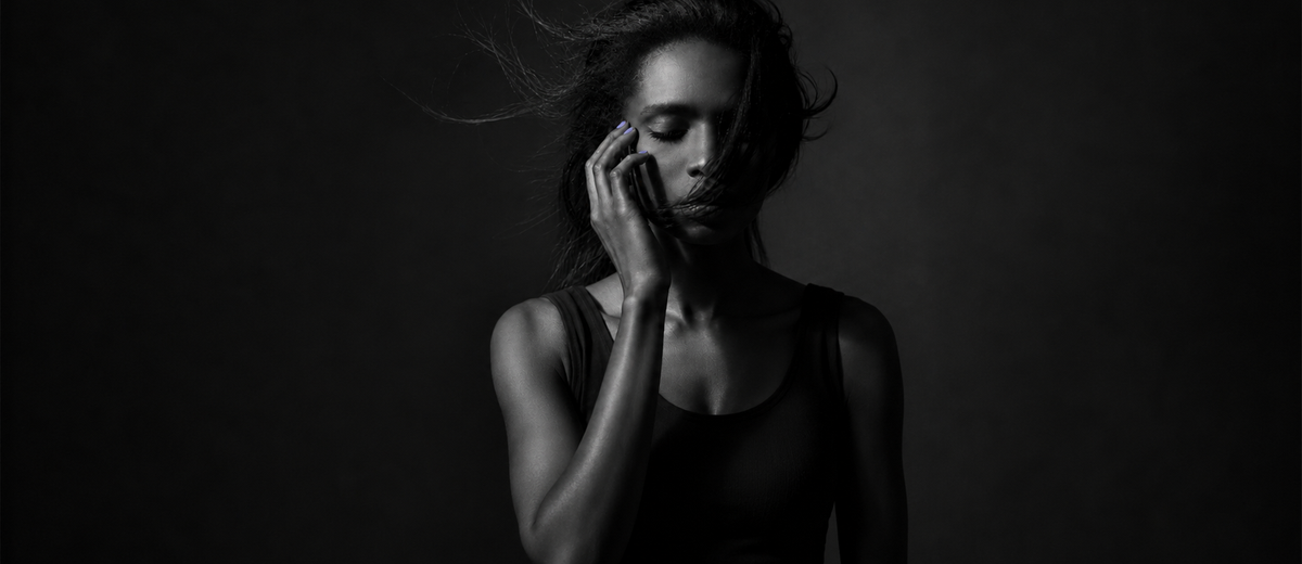 A woman alone in dim light with her eyes closed, sitting with the question of whether infidelity is a dealbreaker.