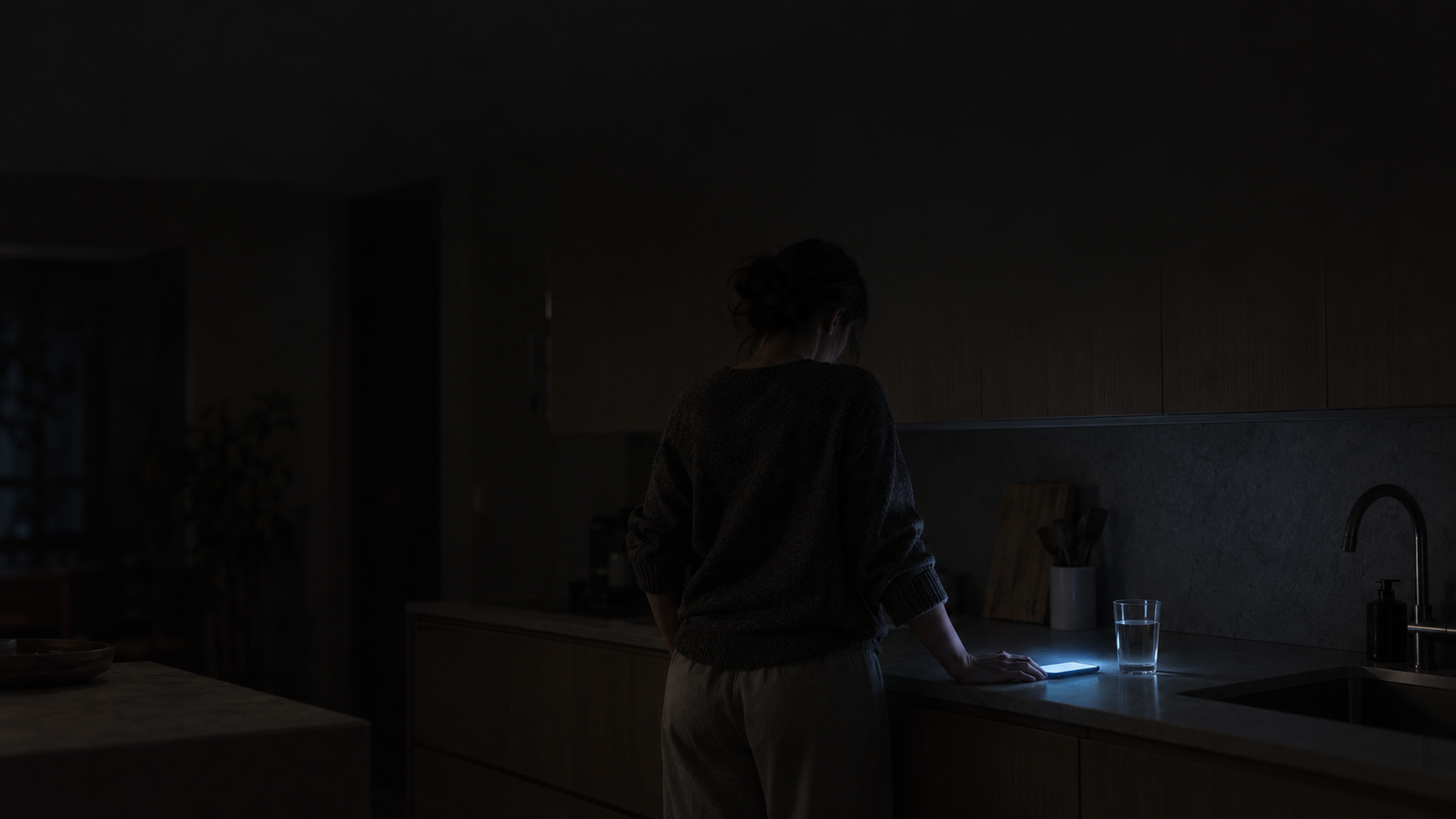 Woman awake alone in a dark kitchen with a phone glowing on the counter, illustrating the checking pattern described in relationship OCD.