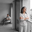 Woman looks away on a balcony as her partner reads apart from her, a pale blue earring the only color in the gray scene