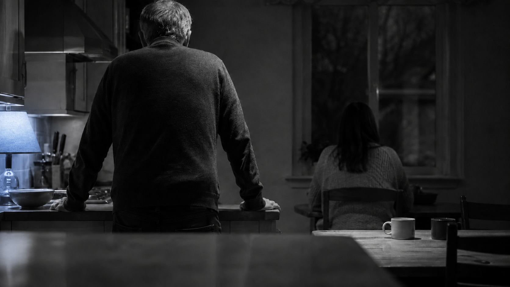 Older man and younger woman apart in a dim kitchen at night, illustrating the distance in age gap relationships