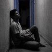 A man sitting alone in a dark hallway at night, reflecting how to regain trust after being cheated on, with pale blue light in the doorway behind him.