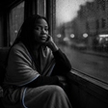 A woman wrapped in a blanket sits alone by a rain-streaked window at dusk, chin resting on her hand, quietly wondering whether her relationship can survive infidelity.
