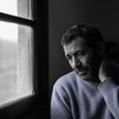 An older man in a pale blue sweater sits alone by a rain-flecked window, head lowered and one hand resting on the back of his neck, his face heavy with quiet exhaustion.