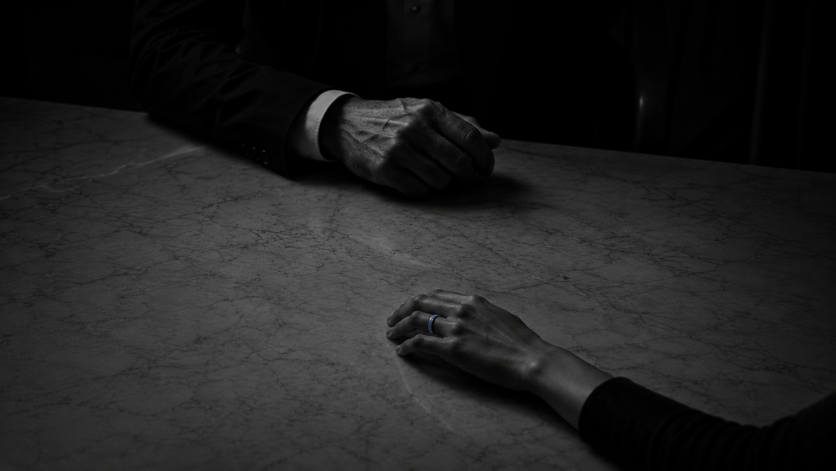 Two hands rest apart on a marble table in dim light. The nearer hand wears a slim blue ring; the other, blurred behind, belongs to someone in a dark suit.