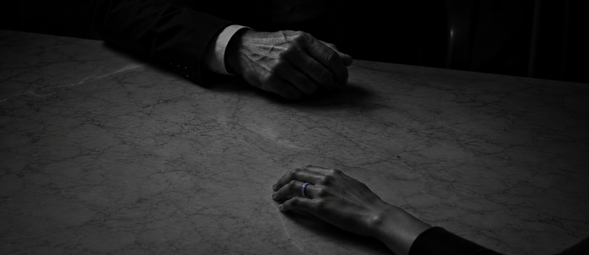 Two hands rest apart on a marble table in dim light. The nearer hand wears a slim blue ring; the other, blurred behind, belongs to someone in a dark suit.