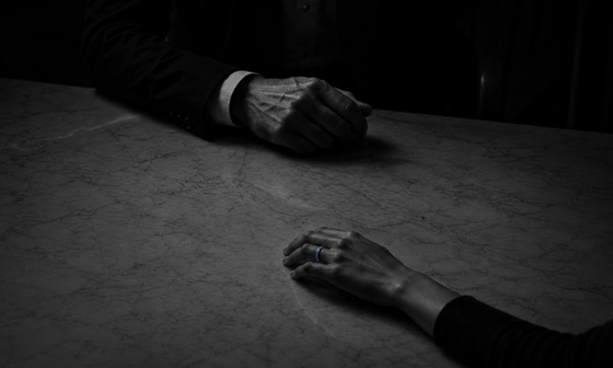 Two hands rest apart on a marble table in dim light. The nearer hand wears a slim blue ring; the other, blurred behind, belongs to someone in a dark suit.