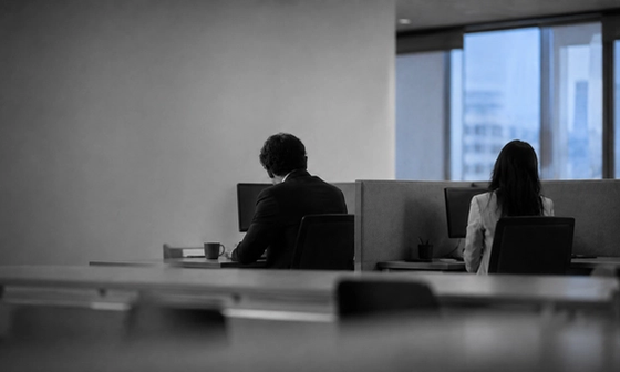Two coworkers seated at opposite ends of a office table, backs to camera, showing the distance of a workplace affair