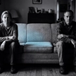 A married couple sits at opposite ends of a couch with an empty cushion between them, evoking the quiet distance of a sexless marriage.