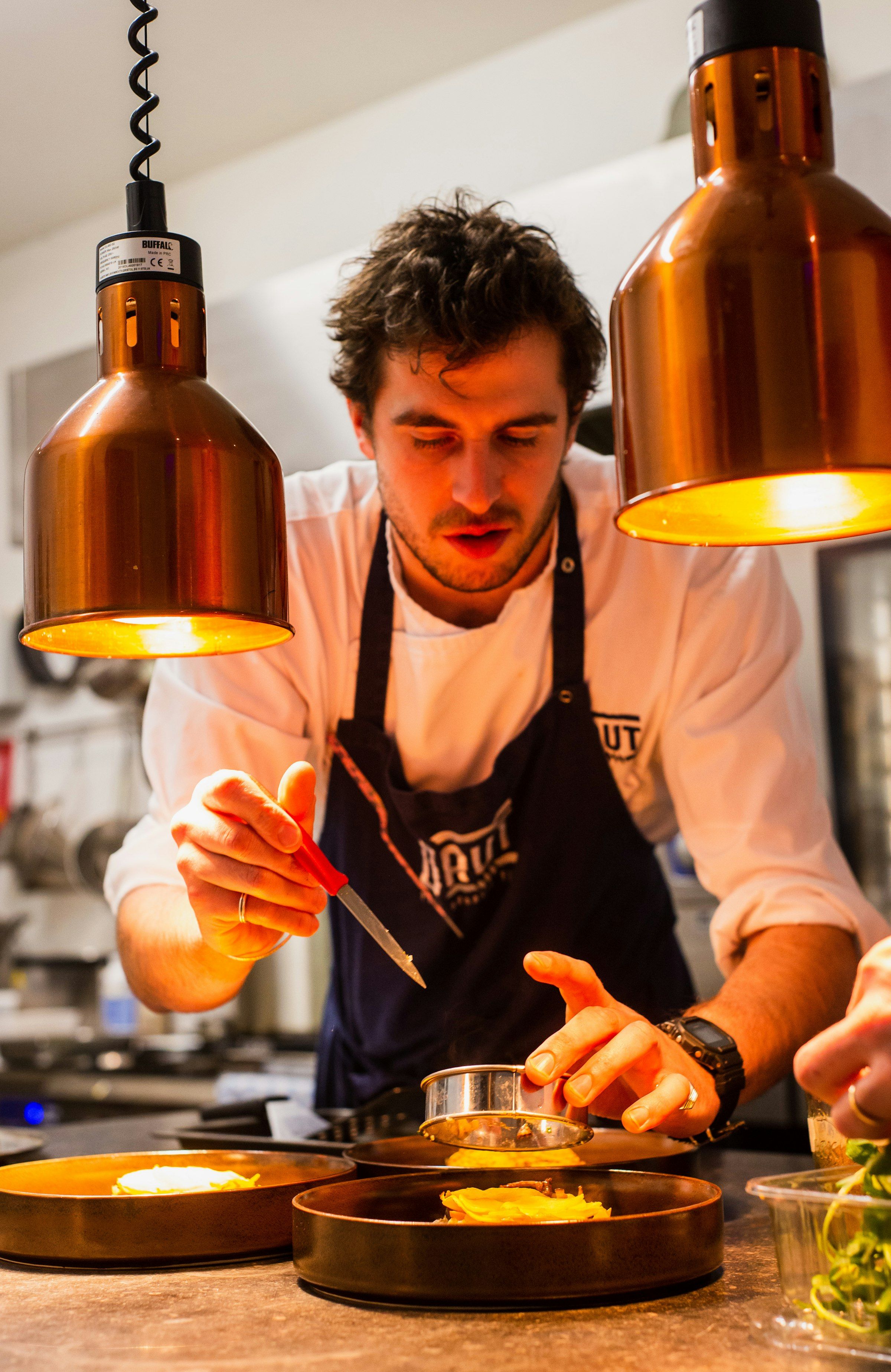 Le chef, tablier noir, dresse une assiette à la pince sous les lampes en cuivre