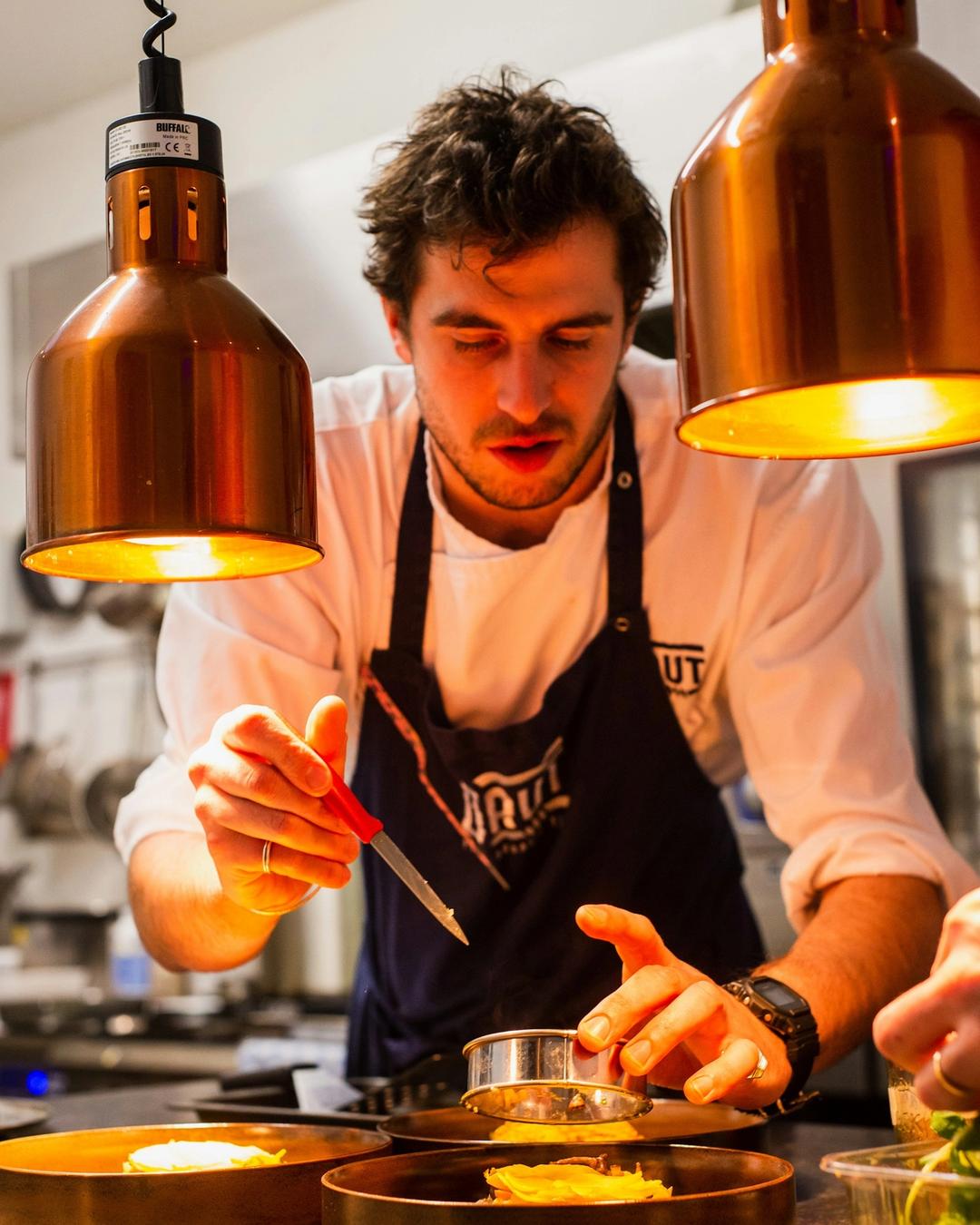 Le chef, tablier noir, dresse une assiette à la pince sous les lampes en cuivre