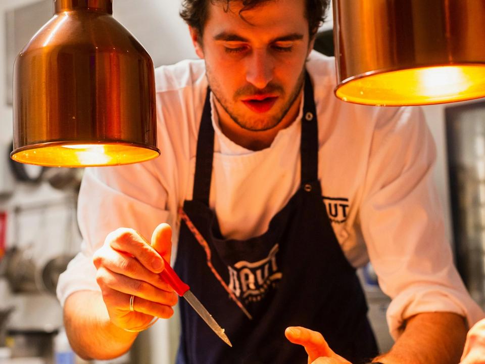 Le chef, tablier noir, dresse une assiette à la pince sous les lampes en cuivre