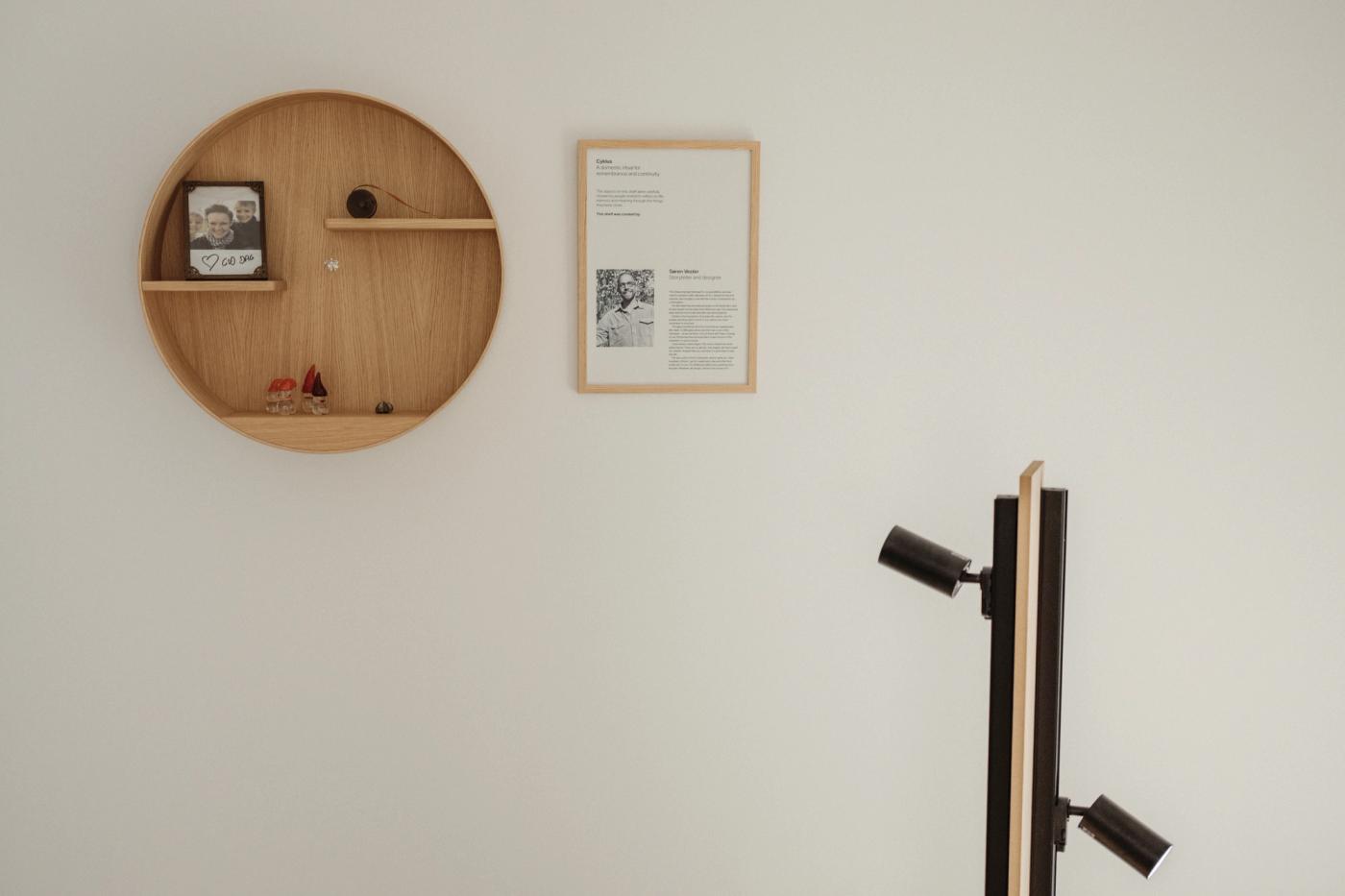 Cyklus memorial shelf in oak, a round wall shelf holding a framed photograph and small objects, beside a framed text panel and a black and oak lamp with two spotlights.