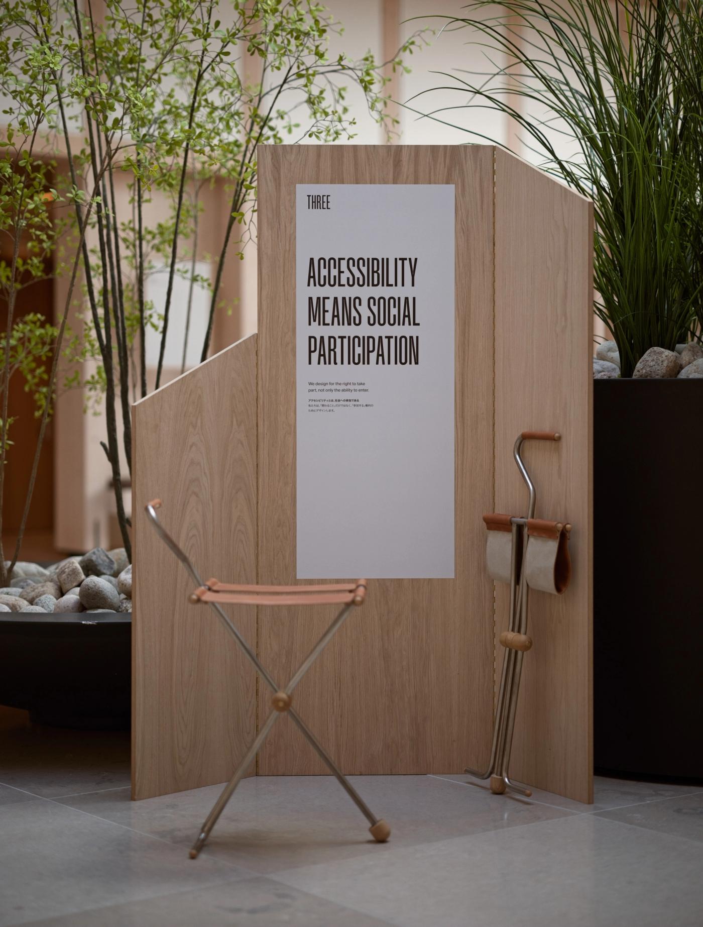 Arch seat cane in oak, stainless steel and leather, with a folding leather seat, beside a stand holding a leather-gripped cane and the poster "Accessibility Means Social Participation".