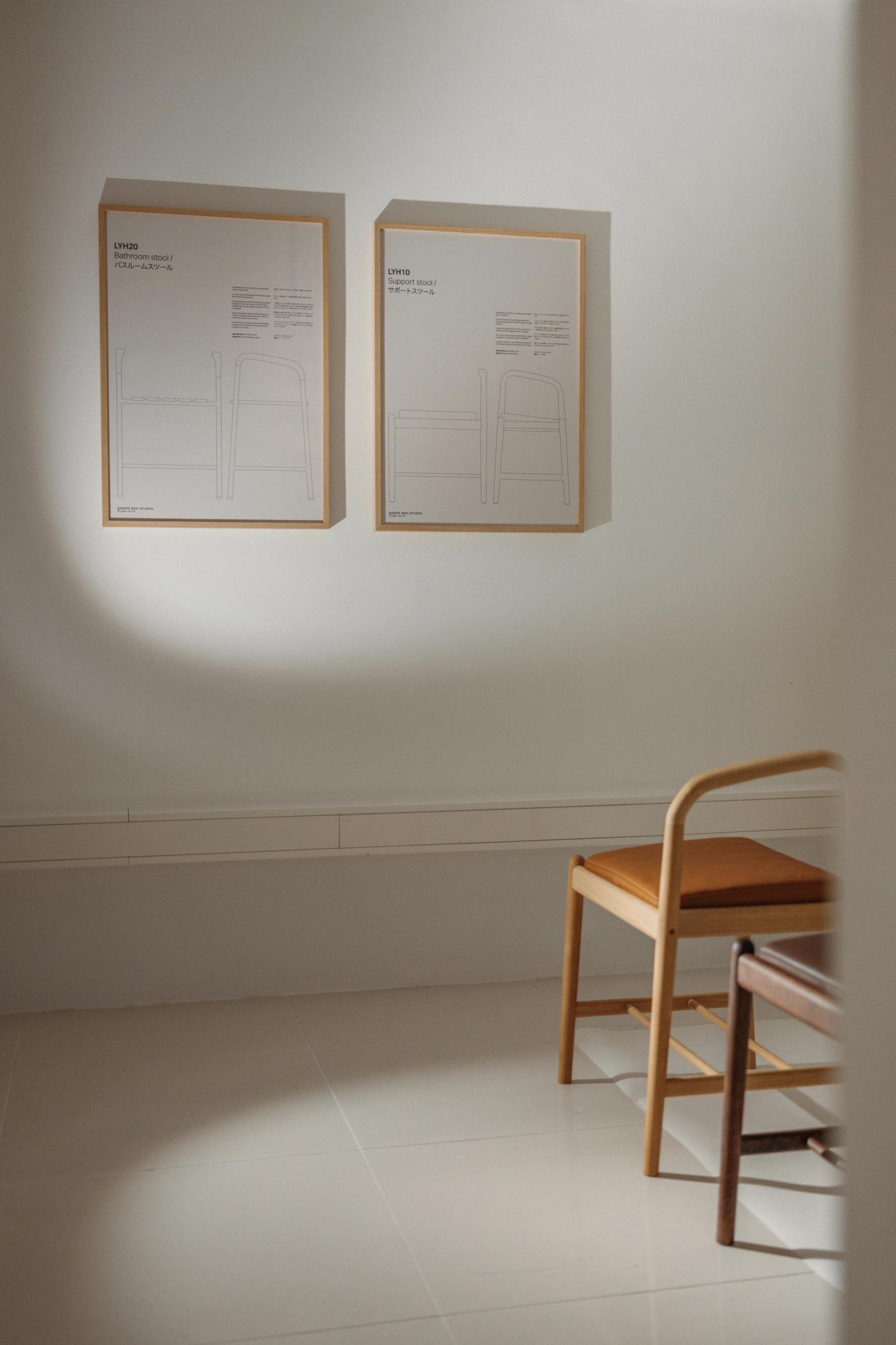 Two framed technical drawings of an arched form on a white wall, beside a wooden chair with a curved backrest and a tan leather seat in soft daylight.