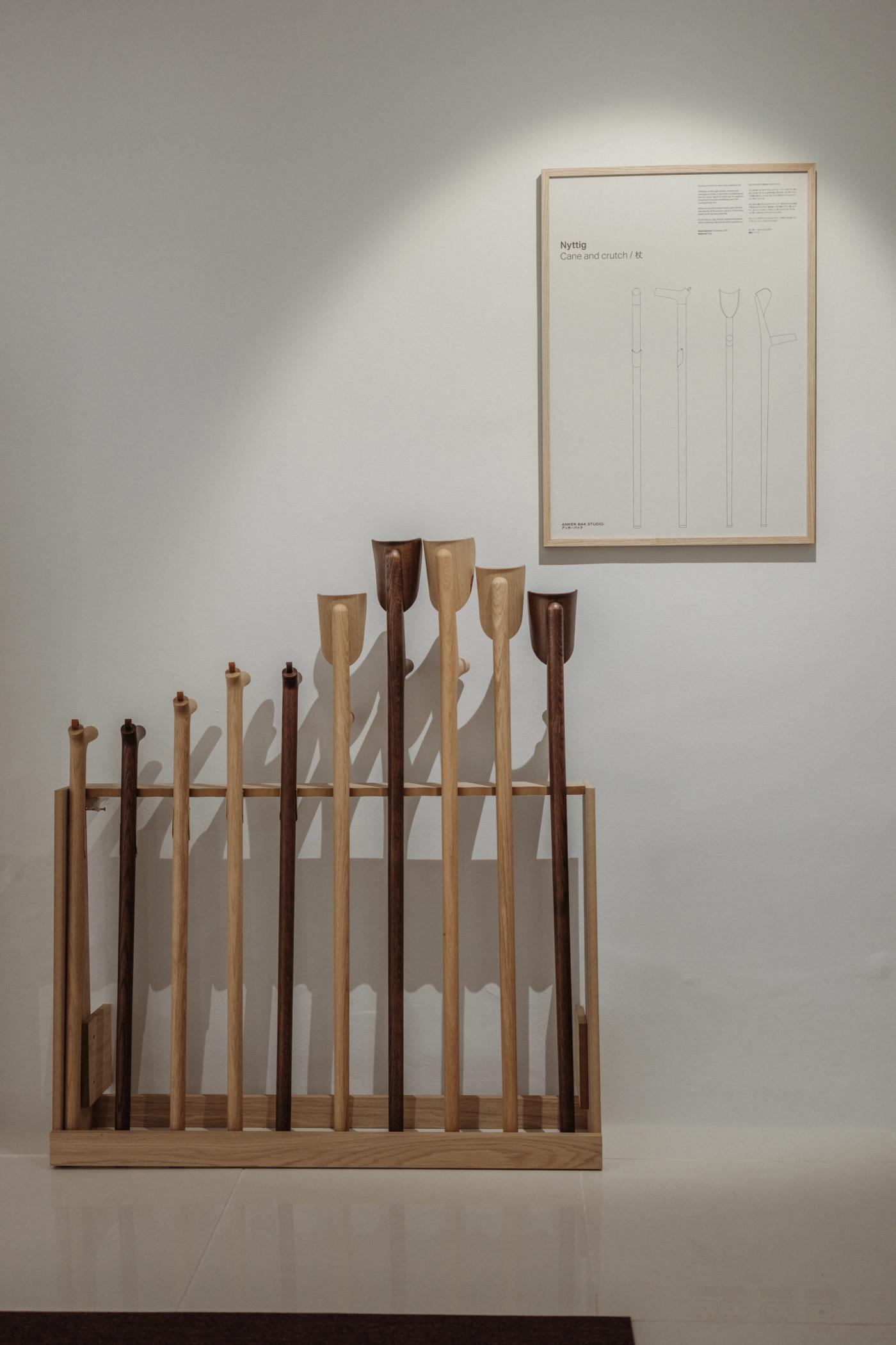 Wooden walking sticks and crutches in light and dark wood of different heights, standing in an oak rack beside a framed drawing on a white wall.