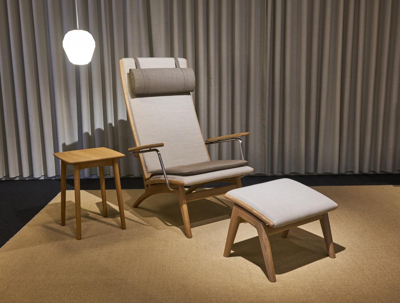 GAZE high-back chair in oak with light upholstery and a neck cushion, beside a matching footstool and a small oak side table on a beige rug.