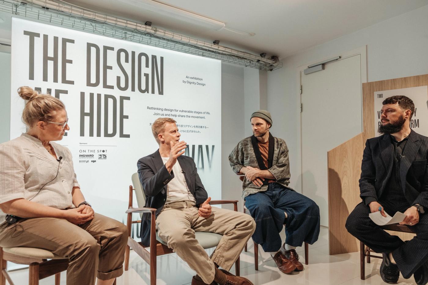 Four people seated on wooden chairs in front of a wall with the exhibition title "The Design We Hide Away", speaking during a panel discussion, with one man gesturing and a man on the right holding notes.