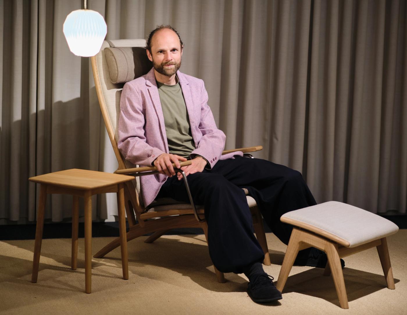 Anker Bak sitting in a GAZE high-back chair with a footstool and a small oak side table, in a pink blazer and dark trousers, beside grey curtains and a white pendant lamp.