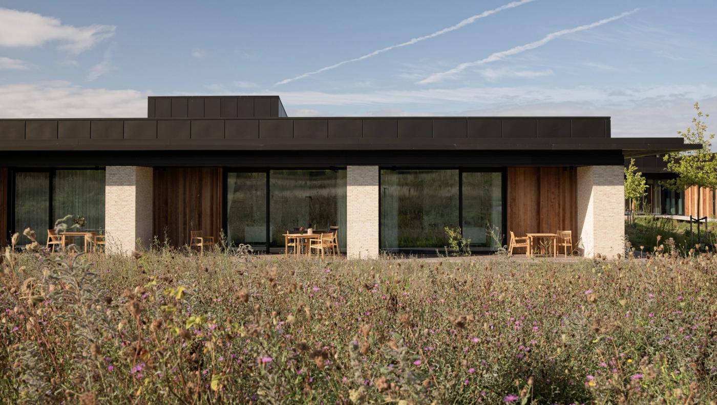 Privathospitalet Mølholm building seen from the garden, with a dark roof edge, timber-clad sections, large windows, light brick pillars and wooden chairs and tables on the terrace behind tall wild grasses.