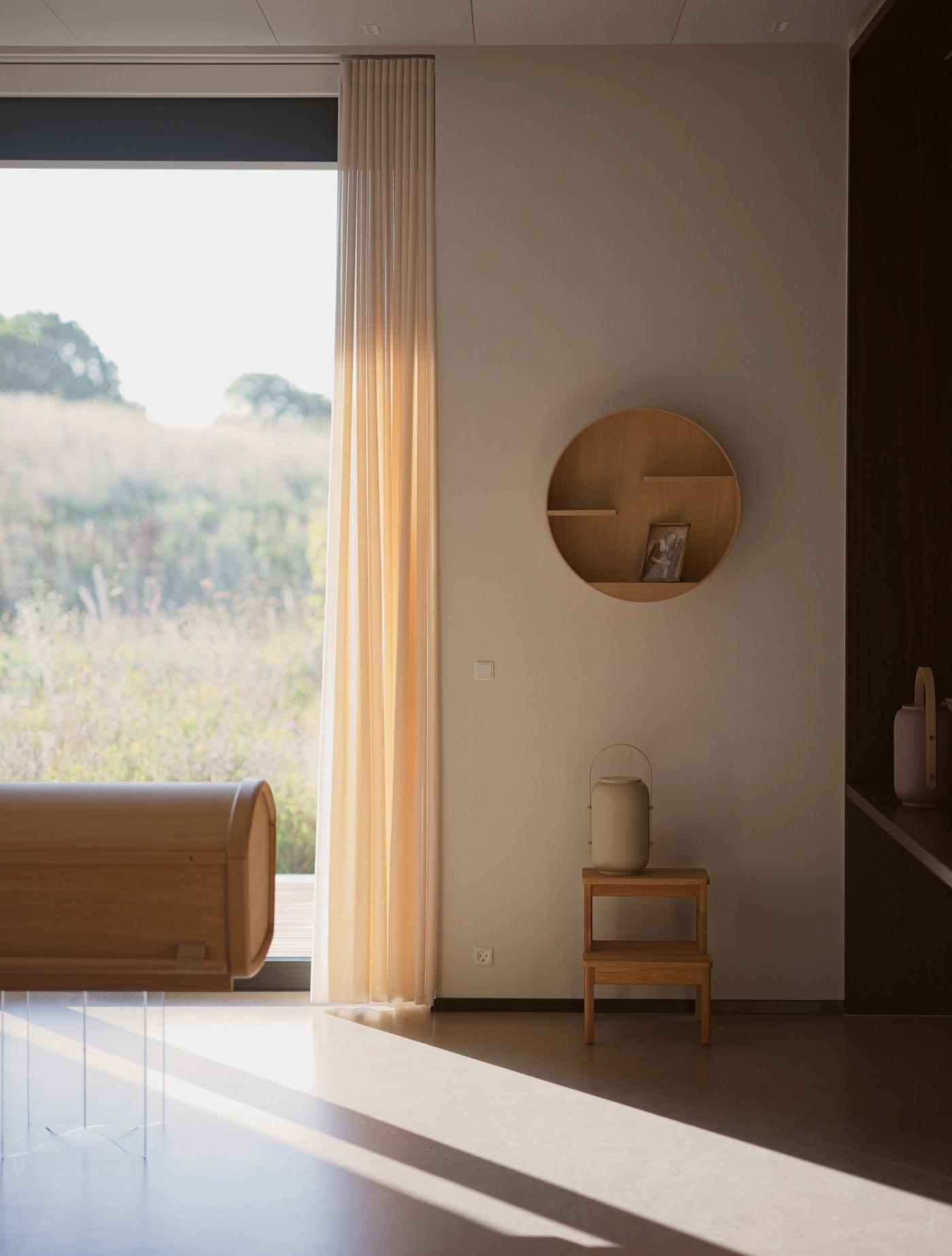 Interior with a round oak memorial shelf and a framed photograph, a cream Akari urn on a small wooden table, and a low wooden form with a rounded end, beside sheer curtains and a window onto a meadow.