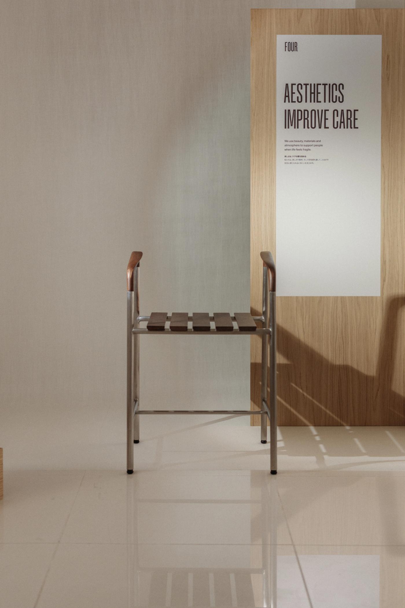 Bath stool with a stainless steel frame, a slatted wooden seat and wooden armrests, in front of an oak panel with the poster "Aesthetics Improve Care".
