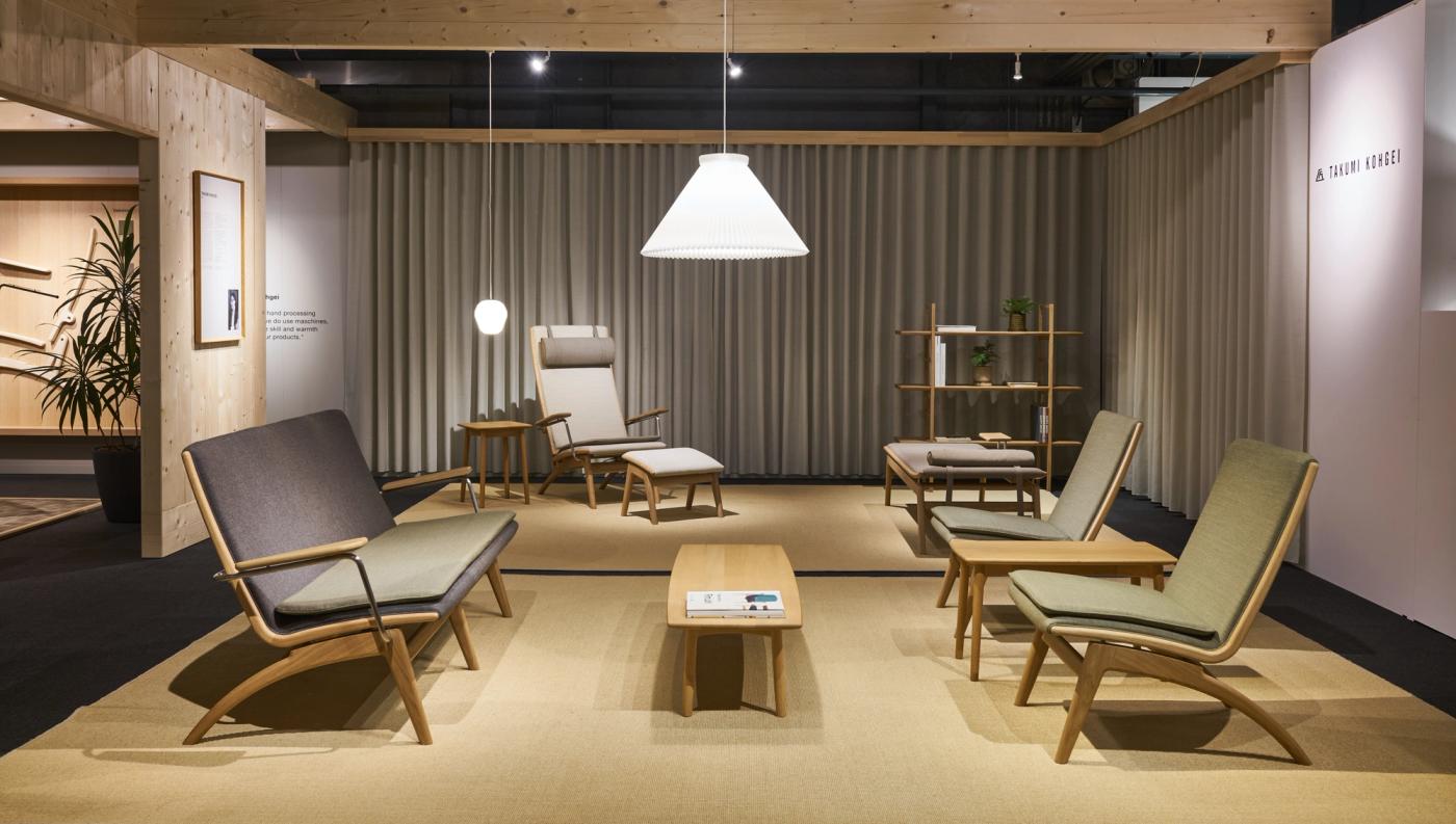 Exhibition room with GAZE lounge chairs and sofa in oak with grey and green upholstery, a GAZE high-back chair with footstool, a GAZE daybed and a low oak table, under a white pendant lamp and beside grey curtains.