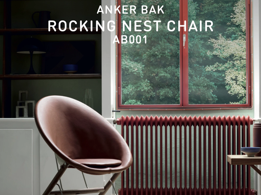 Anker Bak’s Rocking Nest Chair with a rounded upholstered shell and wooden rocking frame, placed on a rug in front of a red radiator and window.
